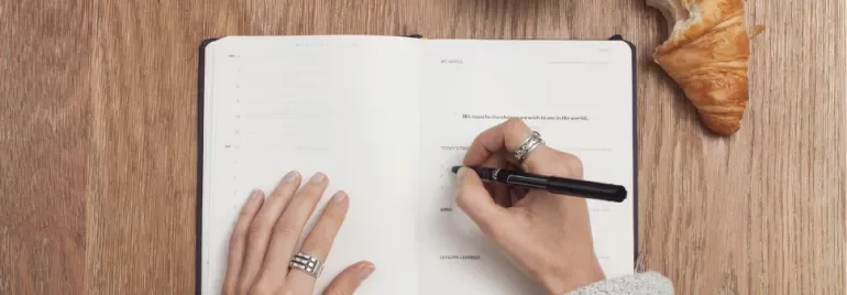 Person writing in a planner next to a croissant on a wooden table.